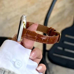 Op de afbeelding: Een zilverkleurige smartwatchbehuizing met een glanzende bruine lederen horlogeband met een gestructureerd krokodillenpatroon. De band is voorzien van een goudkleurige metalen gesp en een bijpassende lederen lus, bevestigd aan de horlogekast.