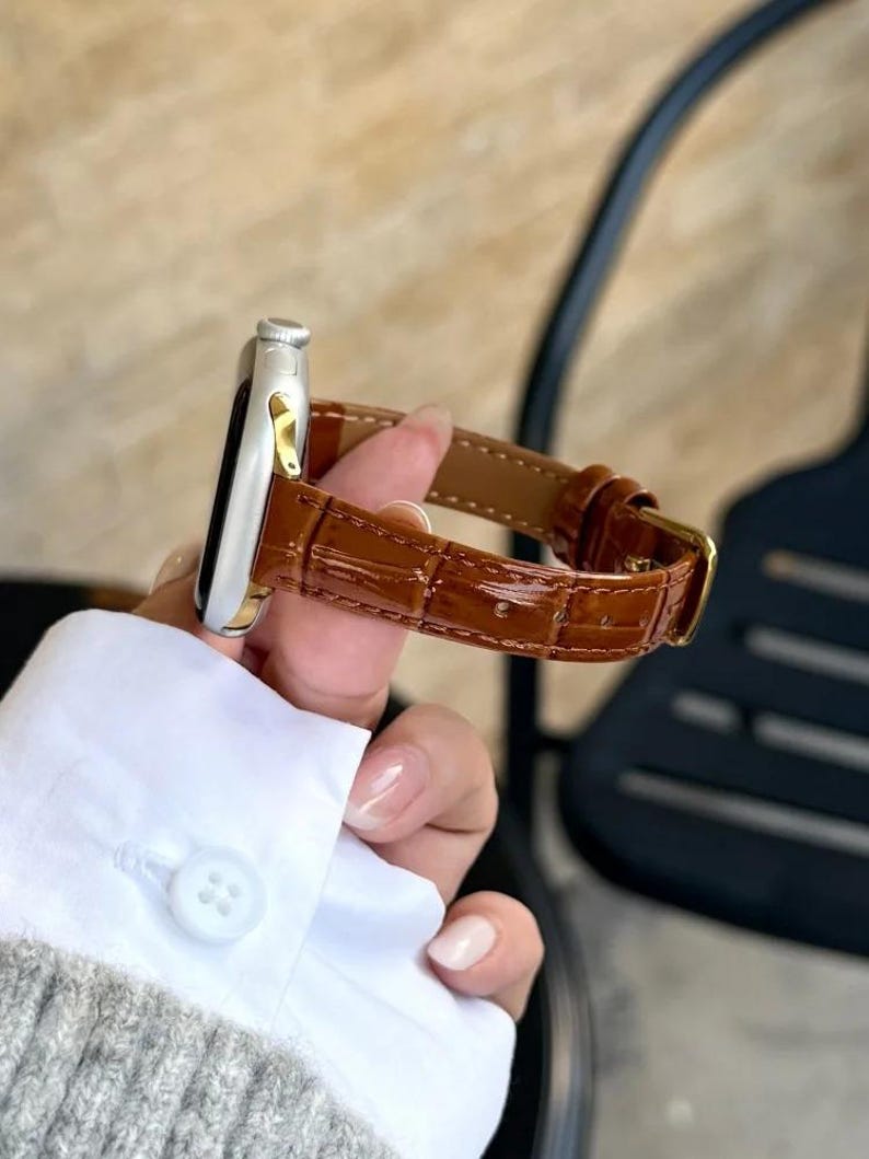 Op de afbeelding: Een zilverkleurige smartwatchbehuizing met een glanzende bruine lederen horlogeband met een gestructureerd krokodillenpatroon. De band is voorzien van een goudkleurige metalen gesp en een bijpassende lederen lus, bevestigd aan de horlogekast.