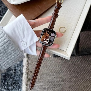 Op de afbeelding: Een smartwatch met een zilverkleurige rechthoekige behuizing en een donkerbruine leren horlogeband met krokodillenprint. Het digitale display toont de tijd 5:12 en een afbeelding van een herfstblad. De horloge heeft een goudkleurige gesp.