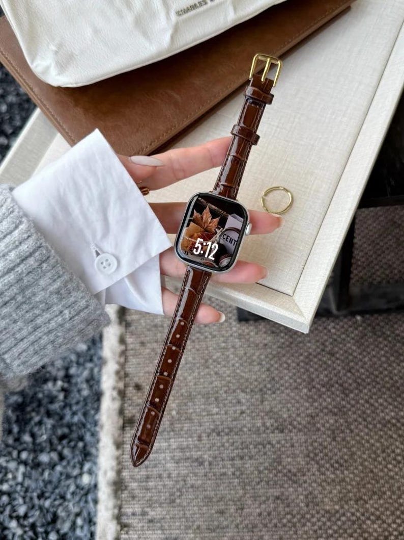 Op de afbeelding: Een smartwatch met een zilverkleurige rechthoekige behuizing en een donkerbruine leren horlogeband met krokodillenprint. Het digitale display toont de tijd 5:12 en een afbeelding van een herfstblad. De horloge heeft een goudkleurige gesp.