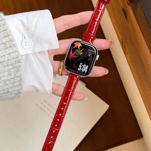 Op de afbeelding: Een smartwatch met een glanzende rode lederen horlogeband met krokodillenmotief en een goudkleurige gesp. Het digitale display toont de tijd 5:04 en een afbeelding van kleurrijke herfstbladeren op een zwarte achtergrond, in een zilverkleurige kast.