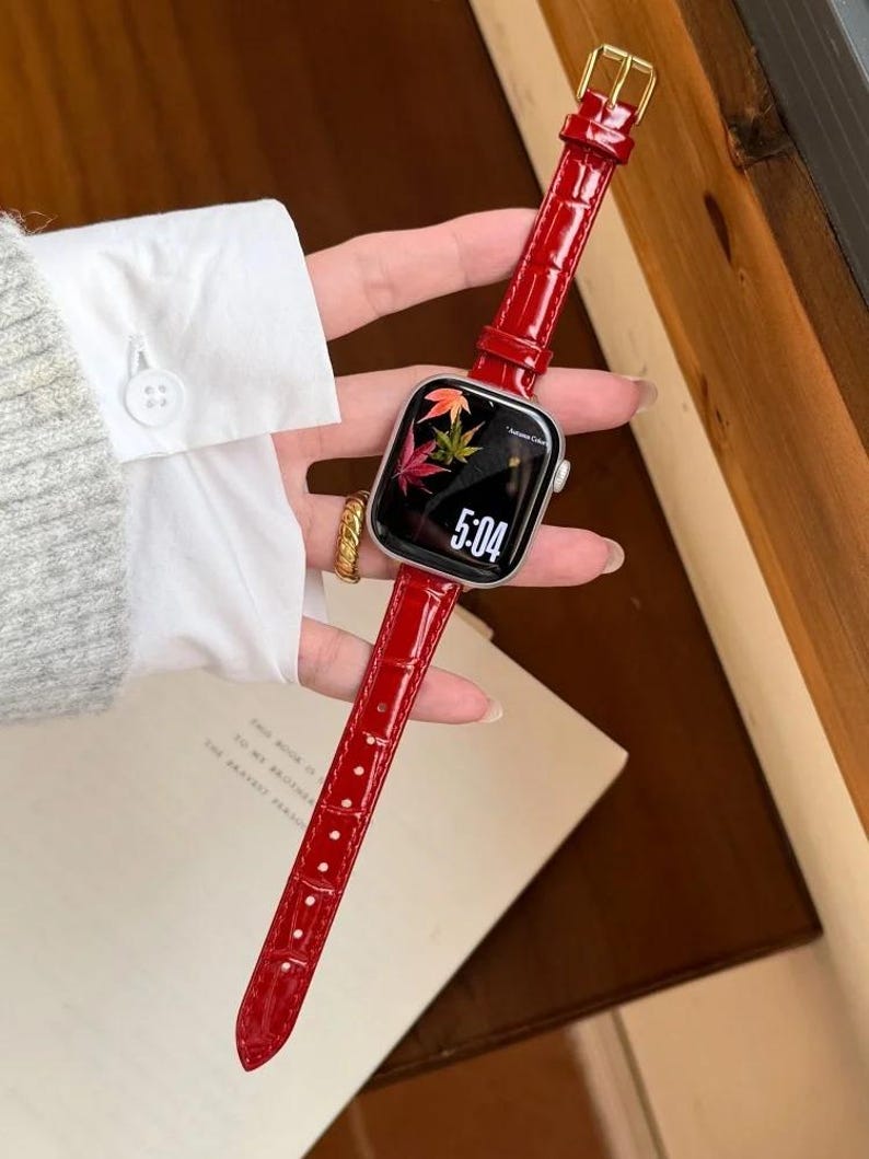 Op de afbeelding: Een smartwatch met een glanzende rode lederen horlogeband met krokodillenmotief en een goudkleurige gesp. Het digitale display toont de tijd 5:04 en een afbeelding van kleurrijke herfstbladeren op een zwarte achtergrond, in een zilverkleurige kast.