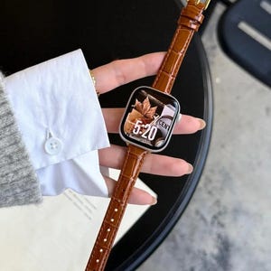 Op de afbeelding: Een smartwatch met een zilverkleurige rechthoekige behuizing en een glanzende bruine leren band met een gestructureerd krokodillenpatroon. Het digitale display toont de tijd 5:20 naast een afbeelding van herfstbladeren en de tekst CENT.