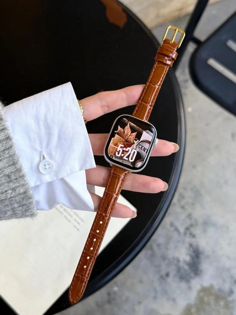 Op de afbeelding: Een smartwatch met een zilverkleurige rechthoekige behuizing en een glanzende bruine leren band met een gestructureerd krokodillenpatroon. Het digitale display toont de tijd 5:20 naast een afbeelding van herfstbladeren en de tekst CENT.