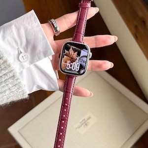 Op de afbeelding: Een smartwatch met een rechthoekige zilveren behuizing en een glanzende, donker magenta lederen horlogeband met een gestructureerd krokodillenpatroon. De wijzerplaat toont een digitale klok die 5:09 aangeeft met een herfstachtige achtergrond.