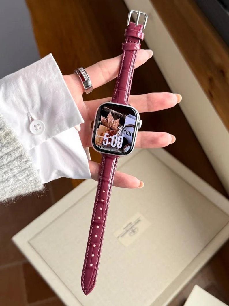 Op de afbeelding: Een smartwatch met een rechthoekige zilveren behuizing en een glanzende, donker magenta lederen horlogeband met een gestructureerd krokodillenpatroon. De wijzerplaat toont een digitale klok die 5:09 aangeeft met een herfstachtige achtergrond.