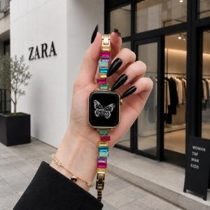 May include: A gold-tone smartwatch with a rectangular face displaying a white butterfly graphic. The watch band features a series of rectangular, multi-coloured crystals in shades of pink, yellow, blue, and green, set in a gold-tone metal link chain.