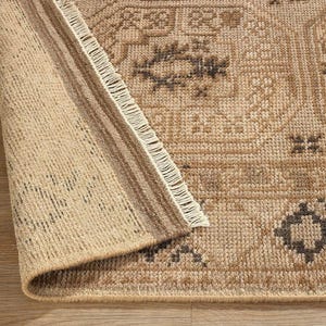 May include: A tan area rug with a folded corner revealing the underside. The rug features a repeating geometric pattern in dark brown and beige, with a visible fringe along the edge of the fold and a textured, woven surface construction.