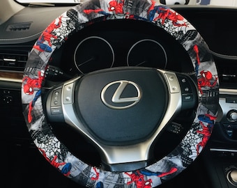 May include: A black steering wheel with a red, white, and gray Spider-Man themed cover. The cover features illustrations of Spider-Man, spiderwebs, and cityscapes. The steering wheel has silver controls and a silver Lexus emblem.