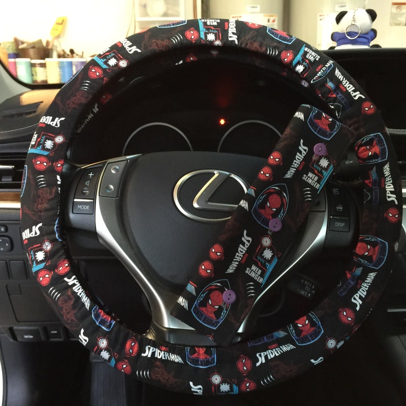 May include: Black steering wheel cover and seatbelt cover with a Spider-Man design. The covers display a repeating pattern of Spider-Man's face and the words "Spider-Man" and "Web Slinger". The covers are on a black steering wheel.