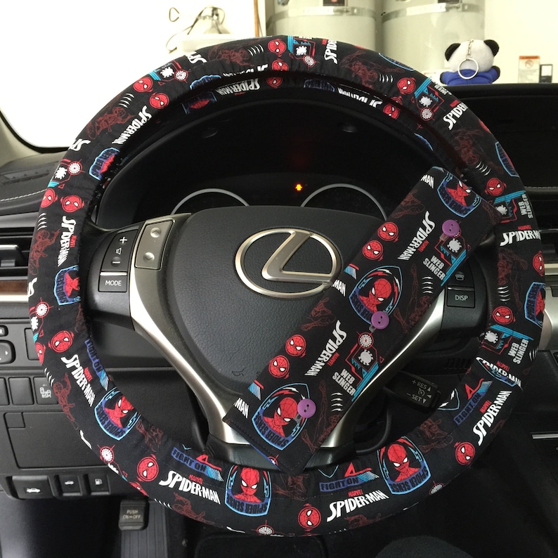 May include: Black steering wheel cover and seatbelt cover set with a Spider-Man design. The covers show Spider-Man graphics, the words "Spider-Man", "Web Slinger", and "Fight On". The steering wheel cover is fitted around a car's steering wheel.