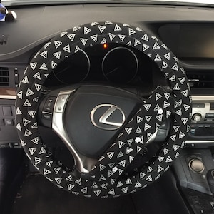 May include: Black steering wheel cover with a white triangle and line pattern. The cover is on a black steering wheel with silver accents. The car's dashboard is visible in the background.