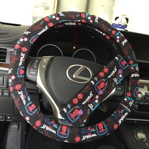 May include: Black steering wheel cover and seatbelt cover set with a Spider-Man design. The covers show Spider-Man graphics and text on a black background. The steering wheel cover is circular, and the seatbelt cover is rectangular.