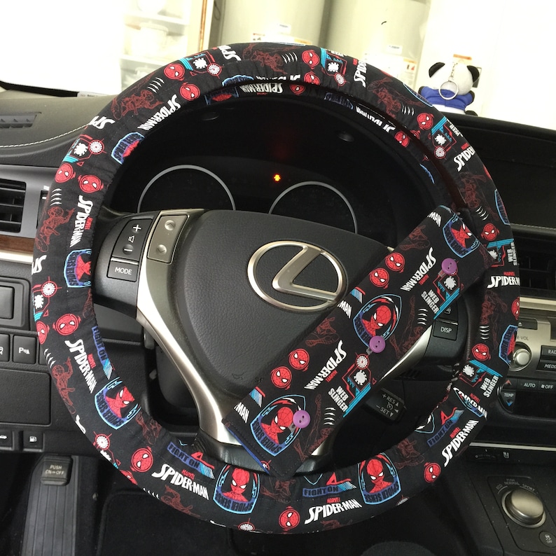 May include: Black steering wheel cover and seatbelt cover set with a Spider-Man design. The covers show Spider-Man graphics and text on a black background. The steering wheel cover is circular, and the seatbelt cover is rectangular.