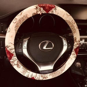 May include: A cream-colored steering wheel cover with a map design and the text "The Marauders". The cover is wrapped around a black steering wheel with silver accents and the Lexus logo.