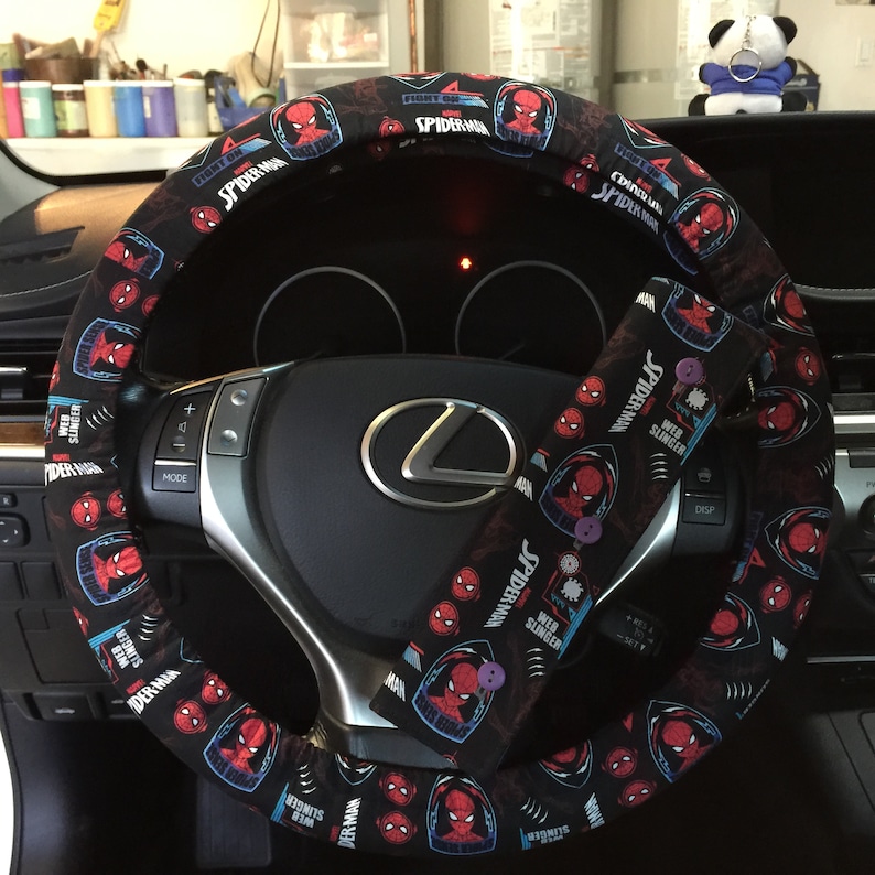 May include: Black steering wheel cover and seatbelt cover with a Spider-Man design. The covers display a repeating pattern of Spider-Man's face and the words "Spider-Man" and "Web Slinger" in white and blue. The steering wheel cover is on a car's steering wheel.