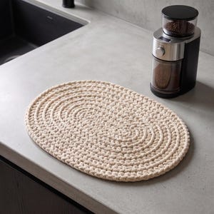 May include: An oval-shaped, cream-colored placemat crafted from thick, braided cord in a continuous spiral pattern. The placemat features a textured, woven surface with visible interlocking loops, providing a sturdy and uniform appearance.