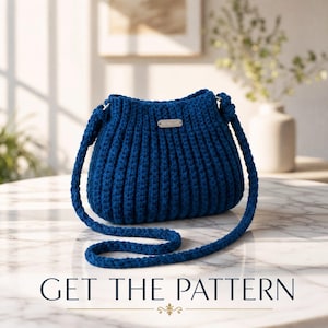 Puede incluir: Un bolso de ganchillo azul intenso con textura acanalada vertical y una correa trenzada a juego. Una pequeña etiqueta metálica rectangular está fijada en el centro. El texto GET THE PATTERN aparece impreso en la parte inferior.
