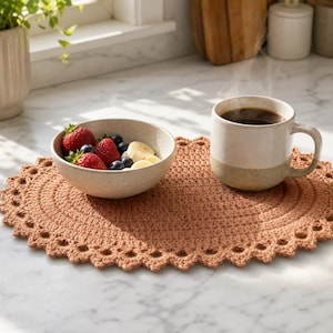 May include: An oval-shaped, terracotta-colored crocheted placemat with a decorative scalloped edge. A light-colored ceramic bowl filled with fresh strawberries, blueberries, and banana slices sits alongside a two-toned ceramic mug of coffee.