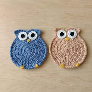 May include: Two round crochet coasters shaped like owls. One coaster is blue and the other is tan. Both feature large white eyes with black pupils, small yellow beaks, and two small yellow feet at the bottom. The crochet pattern is circular.