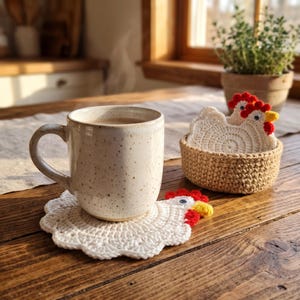 Puede incluir: Una taza de cerámica color crema con motas descansa sobre un posavasos de ganchillo blanco con forma de gallina. Al lado, una pequeña cesta tejida redonda contiene más posavasos de gallina, cada uno con cresta roja, pico amarillo y un ojo móvil.