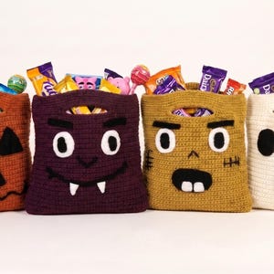 May include: Four rectangular crochet bags in orange, dark purple, mustard yellow, and cream, each featuring a unique face design. The orange bag resembles a jack-o'-lantern, the purple a vampire, the yellow a zombie, and the cream a ghost.