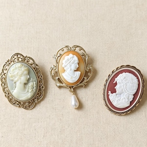 May include: Three oval brooches featuring white relief portraits of profiles. The left brooch has a sage green background and gold filigree frame. The center brooch has an orange background, gold frame, and a dangling white teardrop bead. The right brooch has a maroon background and a twisted gold frame.