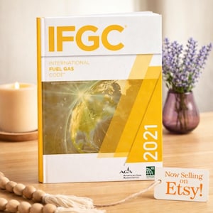 May include: A 2021 International Fuel Gas Code book with a white and yellow cover featuring the text IFGC, the American Gas Association logo, and a graphic of the Earth. A small white tag attached by string reads Now Selling on Etsy.