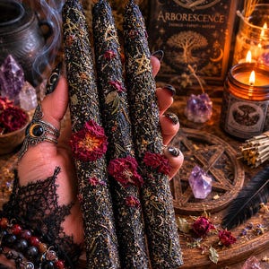 Peut inclure: Trois longues bougies effilées noires décorées de pétales de fleurs rouges séchées, d'herbes vertes et de symboles d'étoiles et de pentagrammes dorés incrustés. Les bougies présentent une surface texturée et rustique.