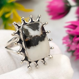 May include: A silver ring featuring a rectangular stone with a white and black marbled pattern. The stone is set in a silver bezel surrounded by a decorative sunburst-style frame with small silver spheres at the end of each radiating prong.