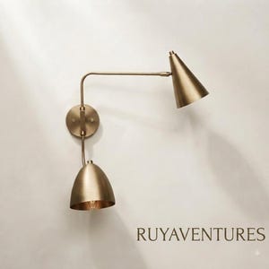 Op de afbeelding: Een wandlamp in een geborstelde messing afwerking met twee verstelbare armen en twee conische kappen. Het armatuur bevat een ronde muurplaat en de tekst RUYAVENTURES staat in de rechterbenedenhoek van de afbeelding gedrukt.