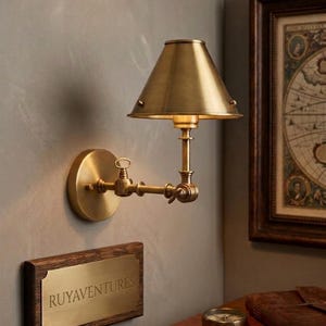 May include: A brass wall-mounted lamp with a conical shade and an adjustable arm. Below the lamp, a rectangular brass plaque mounted on a dark wood frame features the engraved text RUYAVENTURES in a clean, capitalized sans-serif typeface.