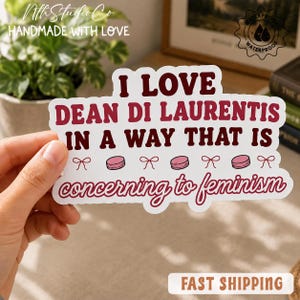 以下が含まれることがあります： I LOVE DEAN DI LAURENTIS IN A WAY THAT IS concerning to feminismという濃い赤色の文字が書かれた白いダイカットステッカー。デザインには、テキストの行の間に小さなピンクのリボンとピンクの円形のアイコンが含まれています。