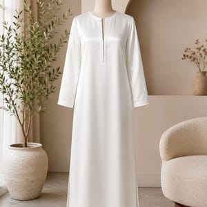 May include: A floor-length, long-sleeved white dress featuring a smooth, satin-like finish, a straight silhouette, and a vertical slit neckline. The garment is displayed on a mannequin, showcasing a clean, solid white color and simple design.