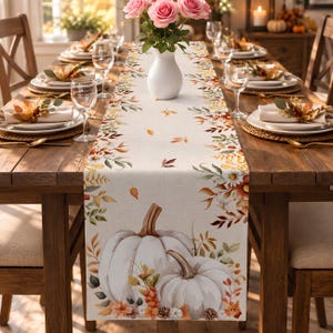 Peut inclure: Un chemin de table rectangulaire orné d'un motif imprimé de citrouilles blanches, de feuilles d'automne, de pommes de pin et de petites fleurs sur un tissu beige clair. Le motif s'étend sur les bords avec des éléments botaniques.