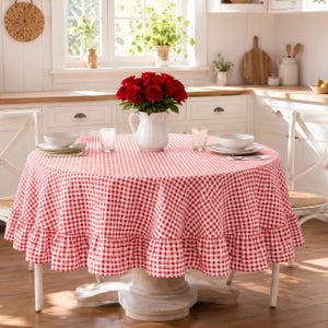 Könnte beinhalten: Eine runde Tischdecke mit rot-weißem Vichy-Karo-Muster und einem gerüschten Rand. Die Tischdecke liegt auf einem runden Tisch, der mit weißen Schüsseln, klaren Gläsern und einem weißen Krug voller roter Rosen gedeckt ist.