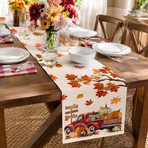 Könnte beinhalten: Ein rechteckiger Tischläufer mit einem Druckmotiv aus verstreuten orangefarbenen und roten Ahornblättern, einem roten Oldtimer-Pickup mit Kürbissen sowie Holzschildern mit dem Text Hello Fall, Give Thanks und Grated auf hellem Grund.
