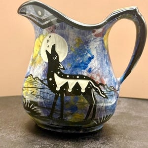 May include: A ceramic pitcher featuring a hand-painted scene of a black wolf howling at a white moon against a blue and yellow mottled background. The pitcher has a curved handle, a pouring spout, and black trim along the rim and handle edges.