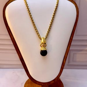 May include: A gold-toned wheat chain necklace featuring a decorative gold-toned pendant with a smooth, round black bead at the base. The pendant has a textured, ornate cap above the bead and a structured bail connecting it to the chain.