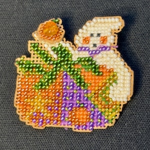 May include: A beaded cross-stitch pin featuring a white ghost with a purple bow, an orange pumpkin with green leaves, and a small orange bead accent. The design is stitched onto a tan perforated base with a scalloped edge and colorful glass beads.