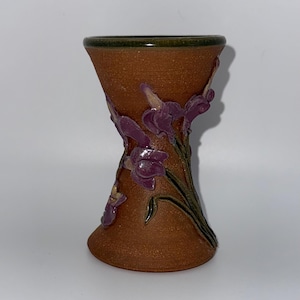 May include: A terracotta-colored ceramic vase with an hourglass shape and a dark green rim. The exterior features raised, three-dimensional purple iris flowers with green stems and leaves applied to the textured, earthy brown clay surface.