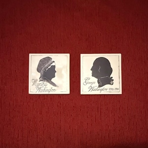 Puede incluir: Dos azulejos de cerámica blancos cuadrados con retratos en silueta negra. El azulejo izquierdo muestra a Martha Washington con su nombre debajo. El derecho muestra a George Washington con su nombre y los años 1732-1799 impresos debajo.