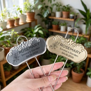 May include: Two metal garden plant markers with a decorative scalloped edge, one silver and one gold. Each features an engraved rose illustration, the text ROSEMARY, Emma's Garden, and the date 04.18.2026, attached to a thin wire stake.