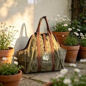 May include: An olive green canvas tote bag with brown leather-like straps and a central pocket. The pocket features a graphic of gardening tools and gloves with the text "Olivia's Garden" printed in white script below the illustration.
