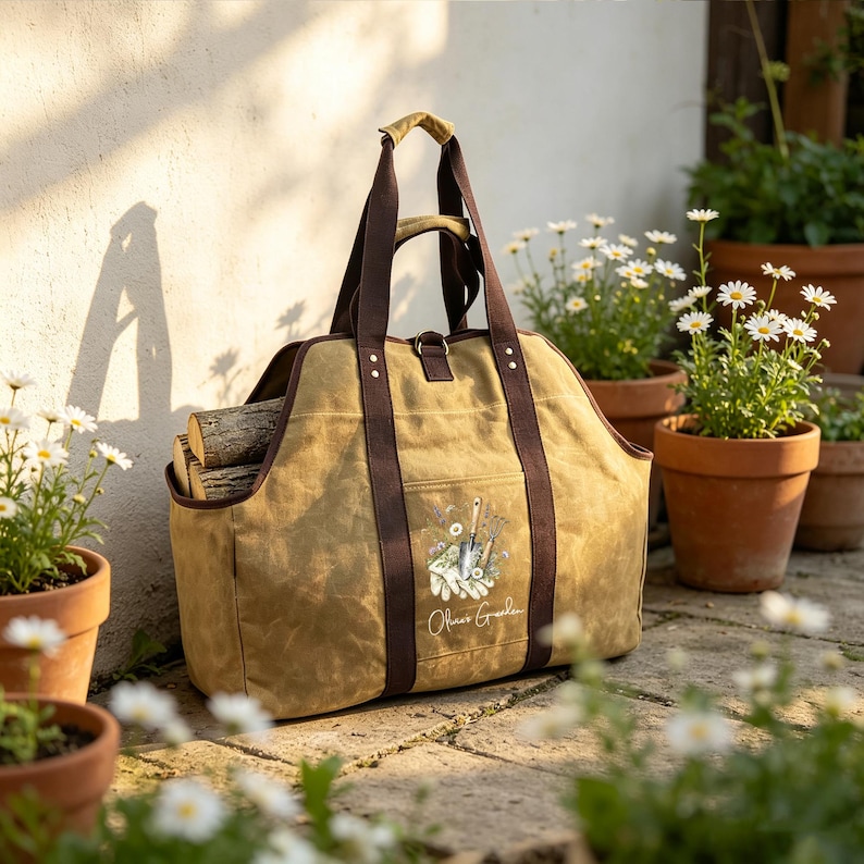 May include: A tan canvas log carrier with dark brown straps and handles. The bag features a front pocket with a printed graphic of gardening tools, gloves, and flowers, along with the text "Olive's Garden". The bag is filled with firewood.