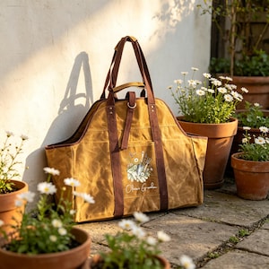 May include: A tan canvas tote bag with brown straps and a central graphic featuring gardening tools, flowers, and the text "Olivia's Garden". The bag has an open top, a structured base, and a vertical strap with a metal ring closure on the front.