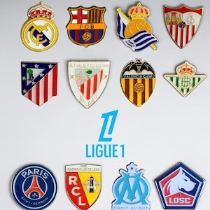 Pode incluir: Uma coleção de doze pins de esmalte metálico com vários logotipos de clubes de futebol profissionais. A seção superior exibe o logotipo da LALIGA EA SPORTS, enquanto a seção inferior apresenta o logotipo da LIGUE 1. Cada pin mostra cores, escudos e símbolos de equipe distintos.