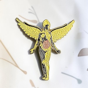 Peut inclure: Une broche en émail métallique représentant une silhouette ailée en position debout. La figure est principalement jaune avec des détails anatomiques noirs et une zone circulaire rose représentant les organes internes au niveau abdominal.