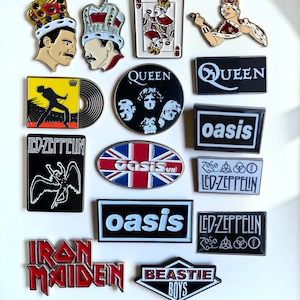 May include: A collection of music-themed enamel pins featuring band logos and portraits. Designs include Queen, Led Zeppelin, Oasis, Iron Maiden, and Beastie Boys. The pins display various shapes, including rectangles, circles, and ovals, with colorful graphics and text.