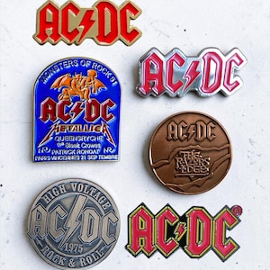 Op de afbeelding: Een verzameling van zes AC/DC metalen emaille pins. De ontwerpen omvatten het klassieke rood-gouden logo, een zilveren logo, een ronde bronzen The Razors Edge pin, een ronde zilveren High Voltage 1975 pin, een rood-zwart logo en een blauwe Monsters of Rock 91 pin.